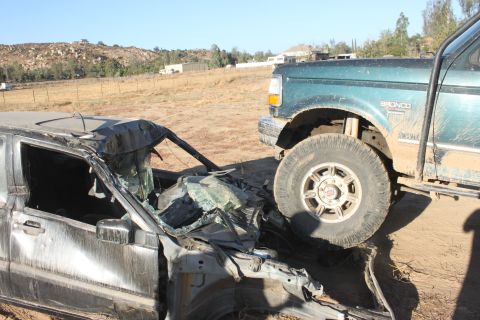 Green 1992 Ford Bronco with "BRONCO" badge on fender, partially crushing a heavily damaged dark sedan
