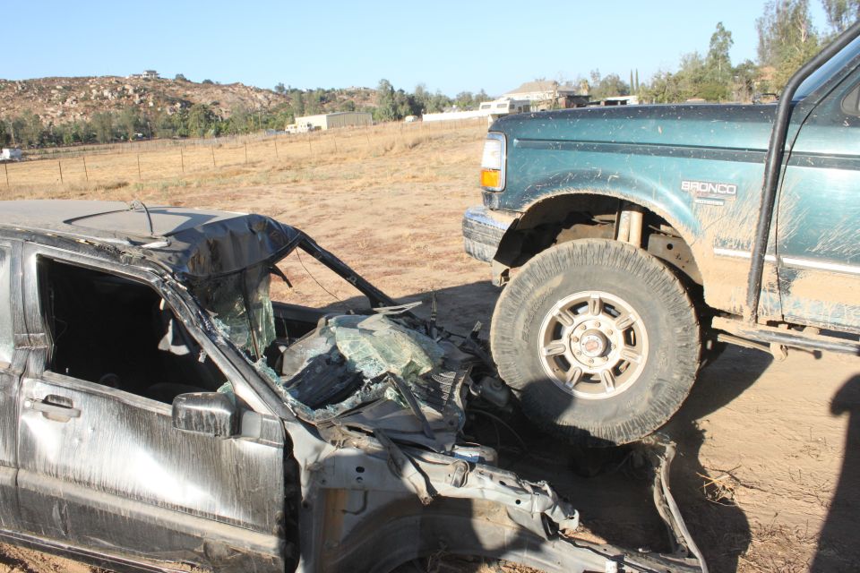 Green 1992 Ford Bronco with "BRONCO" badge on fender, partially crushing a heavily damaged dark sedan