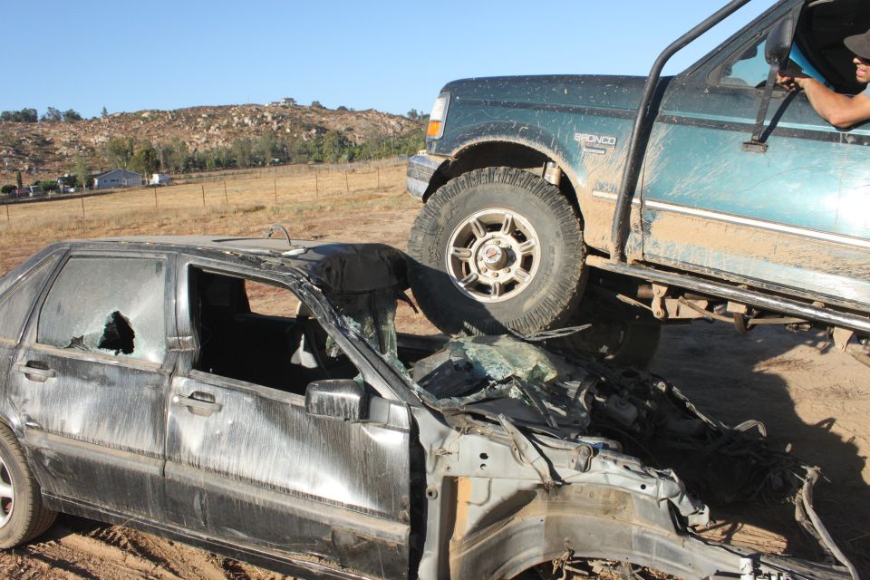 Crushed sedan in front of a green Ford Bronco. Person in driver's seat of Bronco