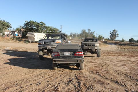 Three lifted trucks in a dusty outdoor area, one in foreground