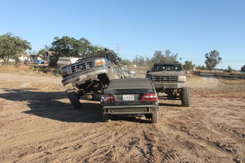 Lifted 1992 Ford Bronco on its side, another lifted Ford truck, and a sedan in a dirt lot