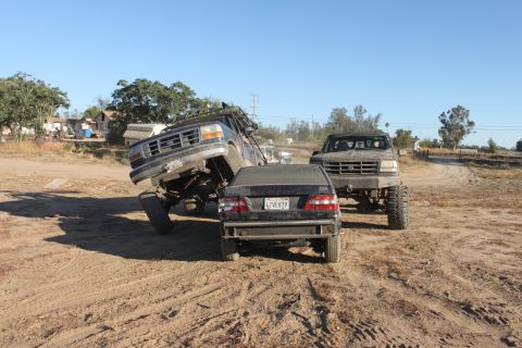 Two lifted trucks are in a dirt lot, one is on top of a sedan. Another truck is behind them