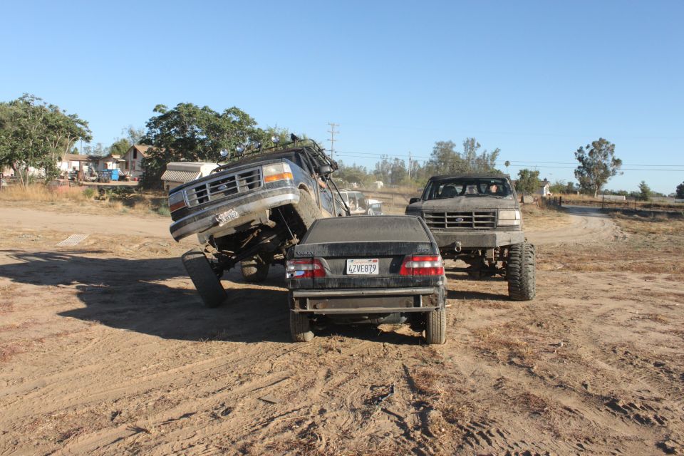 Two lifted trucks are in a dirt lot, one is on top of a sedan. Another truck is behind them