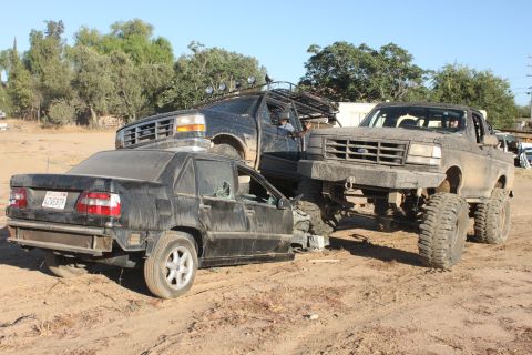 Mud-covered 1992 Ford Bronco on massive tires is parked on top of a damaged black sedan; another black SUV is