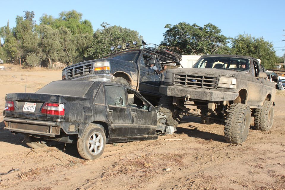 Mud-covered 1992 Ford Bronco on massive tires is parked on top of a damaged black sedan; another black SUV is behind them