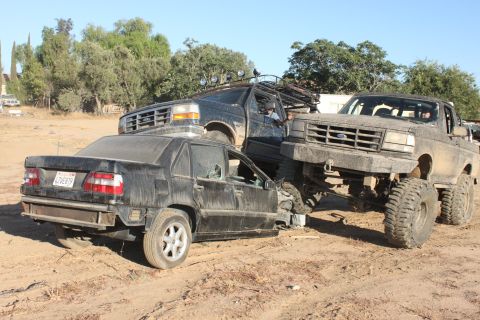 wrecked black sedan, lifted black pickup, and another black truck in a dirt lot