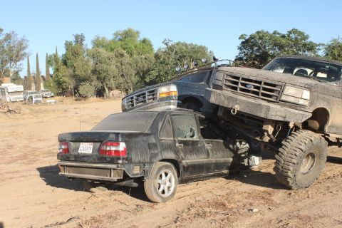 Lifted Ford truck on top of a wrecked sedan, both covered in dust. Truck grille visible