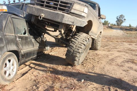 Big Ford truck on top of a black car, both dirty, on a dirt road