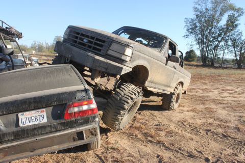 Mud-covered 1992 Ford Bronco on a dirt trail, front grille visible, with a black sedan underneath