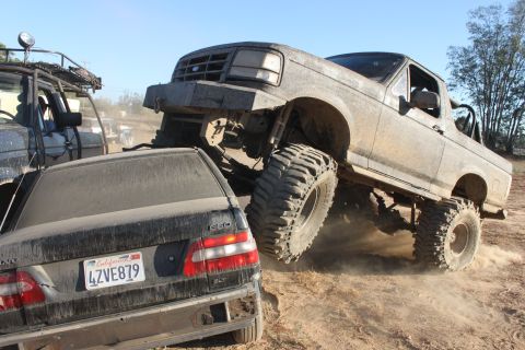 Lifted Ford truck driving over a black Volvo sedan on a dirt trail. Another vehicle is visible to the left