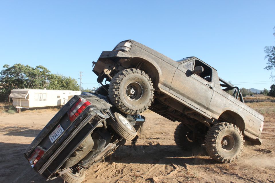 Muddy off-road truck on top of a crushed car, with a trailer in the background