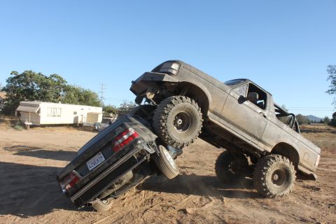 Mud-covered 1992 Ford Bronco on top of a dark sedan, angled upwards on a dirt mound