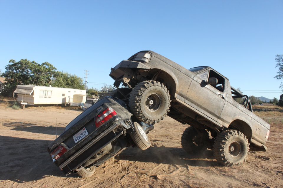 Mud-covered 1992 Ford Bronco on top of a dark sedan, angled upwards on a dirt mound