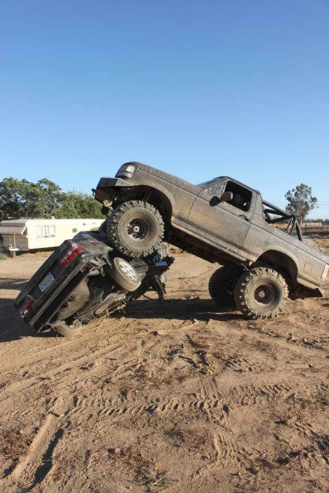 Gray 1992 Ford Bronco on top of a black sedan, both covered in dirt, on a sandy terrain