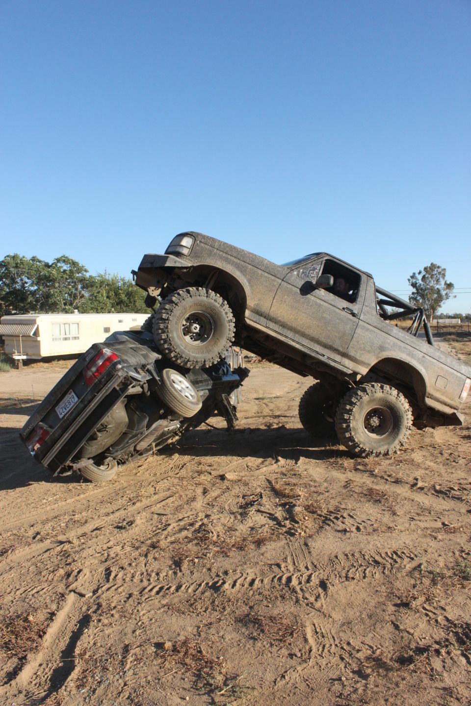 Gray 1992 Ford Bronco on top of a black sedan, both covered in dirt, on a sandy terrain
