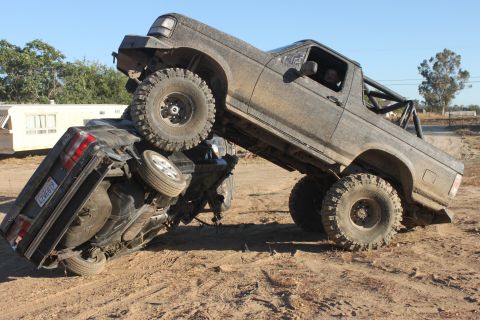 Mud-covered 1992 Ford Bronco on top of an overturned black car, off-road tires, roll cage