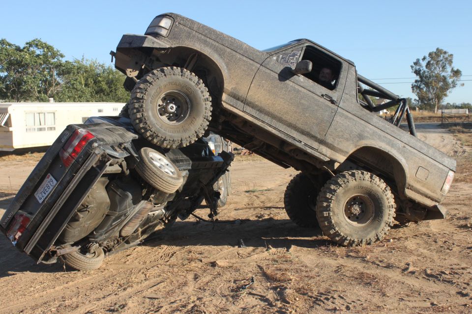 Mud-covered 1992 Ford Bronco on top of an overturned black car, off-road tires, roll cage