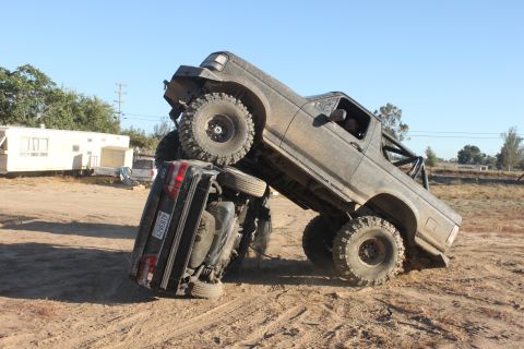 Gray off-road truck on top of a black car, lifted, with large tires