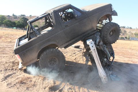 1992 Ford Bronco with roll cage on top of a crushed vehicle, off-road tires, smoke from wheel