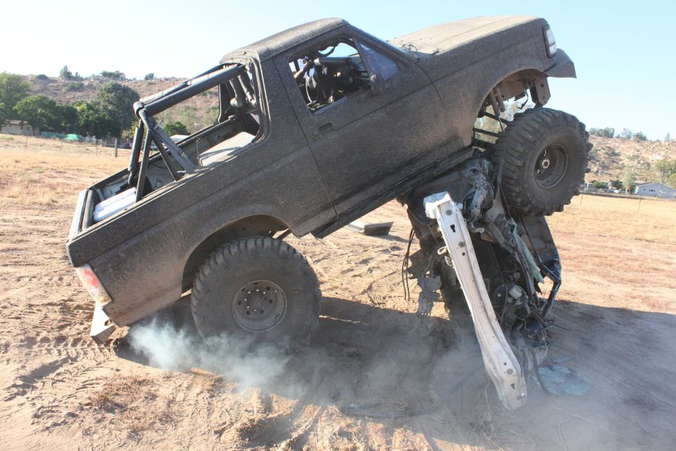 1992 Ford Bronco with roll cage on top of a crushed vehicle, off-road tires, smoke from wheel