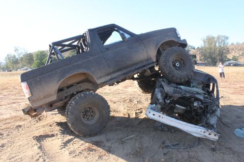 Black off-road Bronco on top of a crushed car, with a roll cage in the back. Person standing in background
