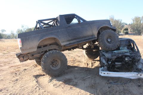Black off-road truck with large tires on top of a crushed car. Roll cage visible in truck bed