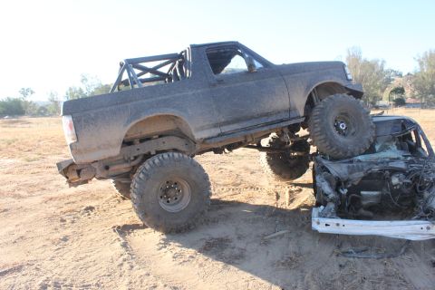 Dark gray 1992 Ford Bronco with a roll cage, lifted suspension, and large tires, crushing a wrecked white car