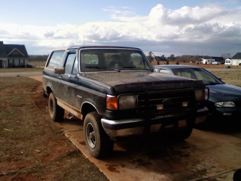 1990 Ford Bronco - A dusty black Ford Bronco sits on a concrete driveway under a cloudy sky, with a sedan parked nearby.