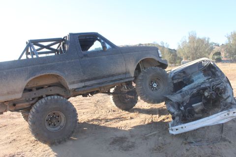 Dark gray 1992 Ford Bronco with large off-road tires crushing a white car