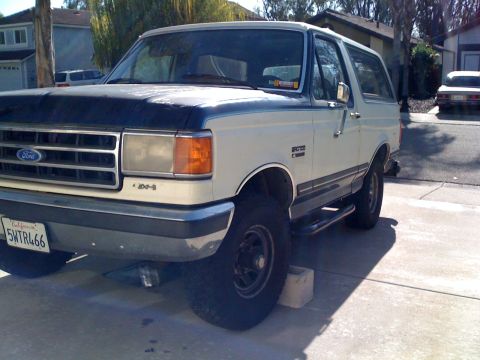 1989 Ford Bronco - A white 1989 Ford Bronco with a dark hood is parked on a driveway, with a residential area in the background.