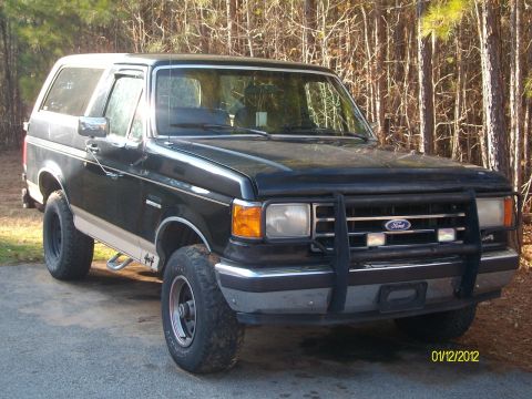 1990 Ford Bronco - This Ford Bronco features a bull bar and off-road tires, parked in a wooded area.
