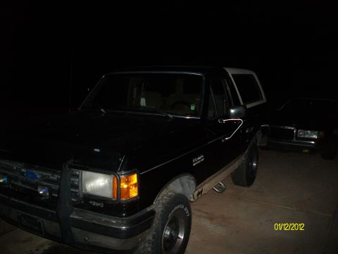 1990 Ford Bronco - A black 1990 Ford Bronco with a white top is seen parked next to another vehicle at night.