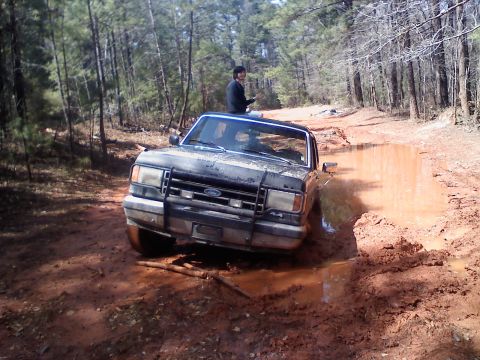 1990 Ford Bronco - A 1990 Ford Bronco navigates a muddy trail, with a person visible through the sunroof.