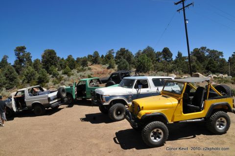 1989 Ford Bronco - A collection of off-road vehicles, including a yellow Jeep and a white Ford Bronco, are parked together on a dirt lot under a clear blue sk…