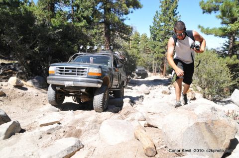 1989 Ford Bronco - A man walks alongside a lifted Ford Bronco as it navigates a rocky off-road path in a wooded area.
