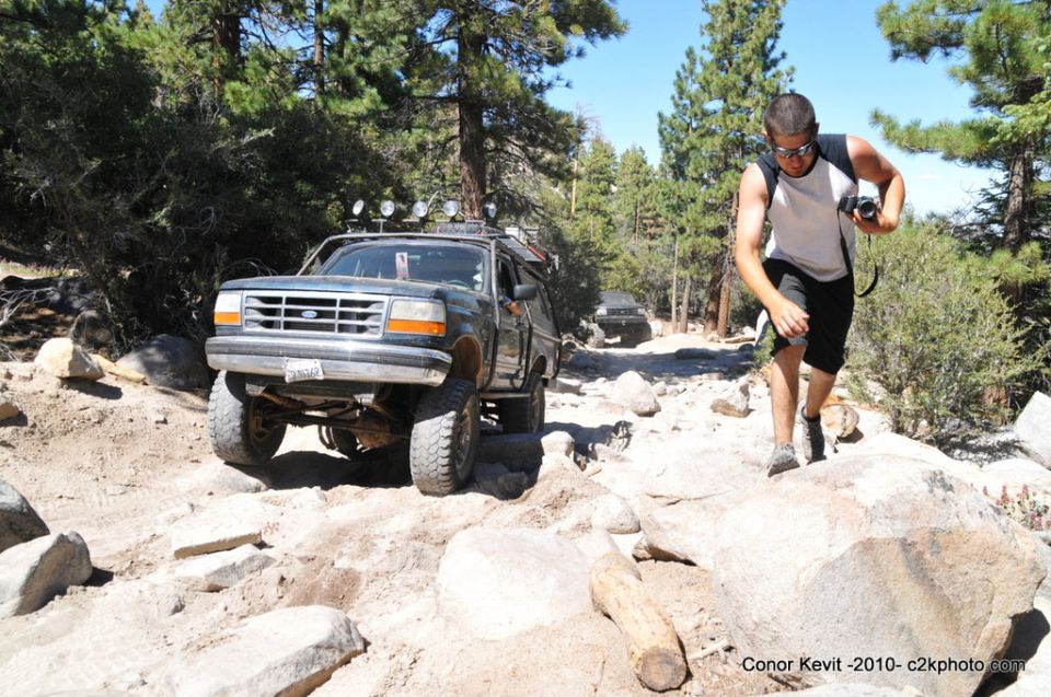 Ford Bronco driving over rocks on a dirt trail, guy with camera walking nearby
