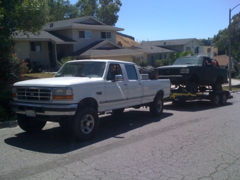 1989 Ford Bronco - A white Ford F-250 tows a black Ford Bronco on a trailer down a suburban street.