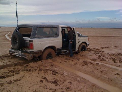 1989 Ford Bronco - This 1989 Ford Bronco is deeply stuck in a muddy, cracked earth landscape with two people visible inside and one standing by the open door.
