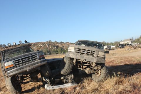 Two lifted Ford trucks, one on top of the other, in a field. The front truck is a 1992 Ford Bronco