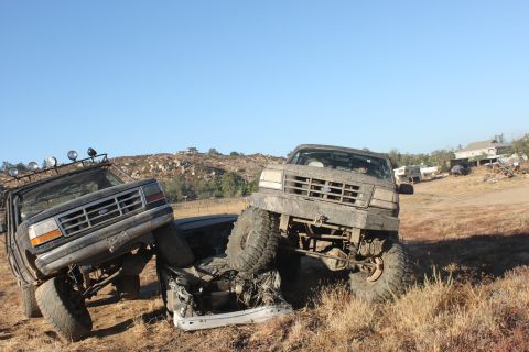 Two muddy Ford trucks are crashed together, one on top of the other, with a clear blue sky overhead