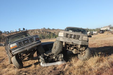 Two lifted Ford trucks, one on top of the other, stuck in dirt. The front truck is a 1992 Ford Bronco