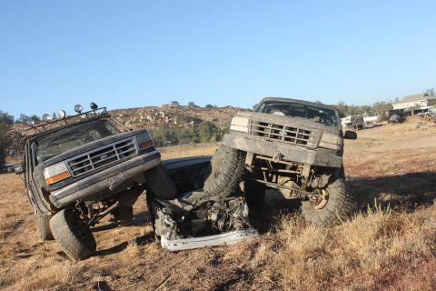 Two lifted Ford trucks are stuck in a field, one on top of a crushed car