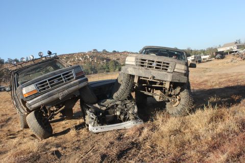 Two muddy Ford Broncos, one on top of a crushed car, the other partially obscuring it