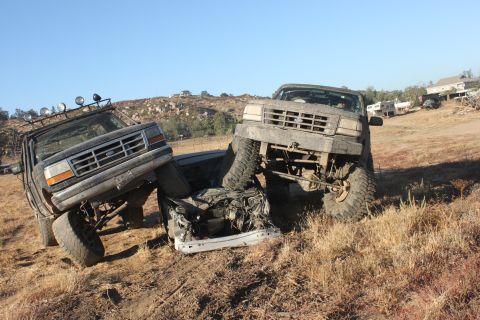 Two mud-covered 1992 Ford Broncos are positioned to crush a white car on a dry, grassy hillside
