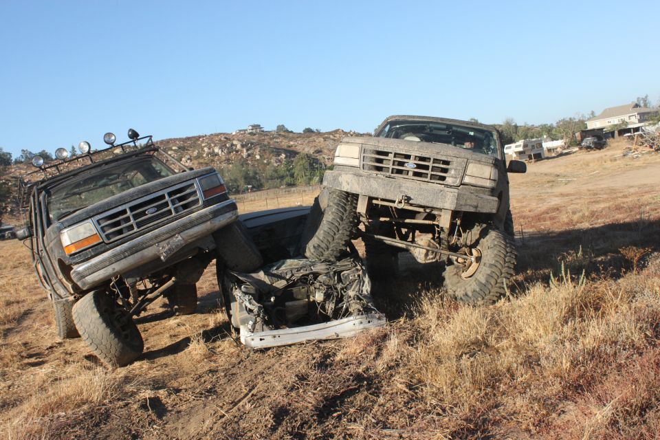 Two mud-covered 1992 Ford Broncos are positioned to crush a white car on a dry, grassy hillside