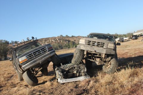 Two Ford trucks are wrecked and stuck in dry grass, one on top of the other