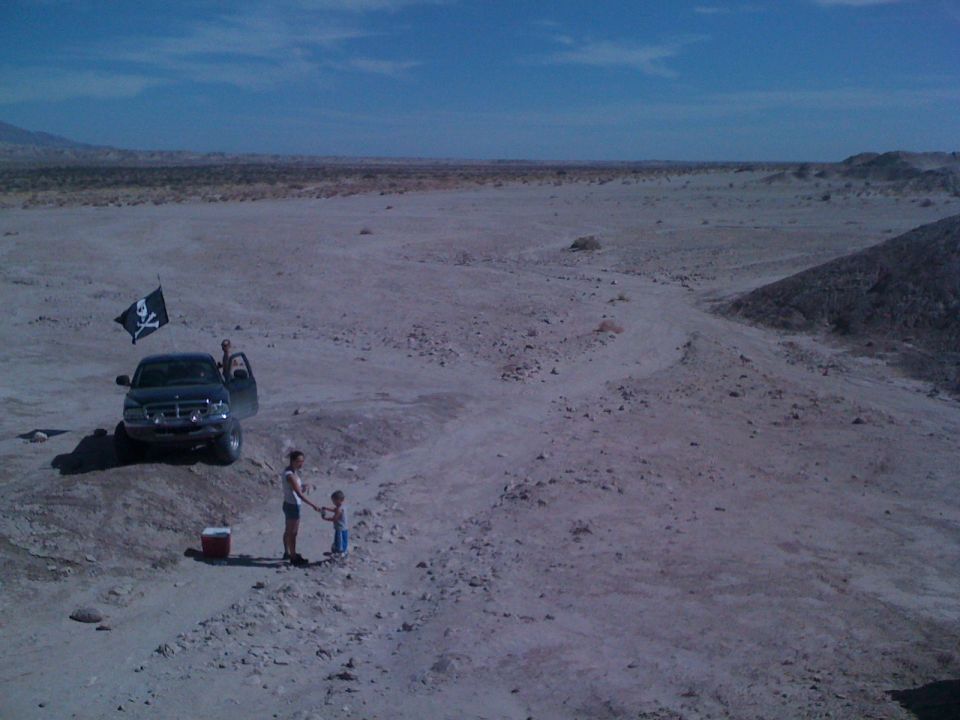 Truck parked in desert with pirate flag flying. Two people stand near it, one holding a child's hand