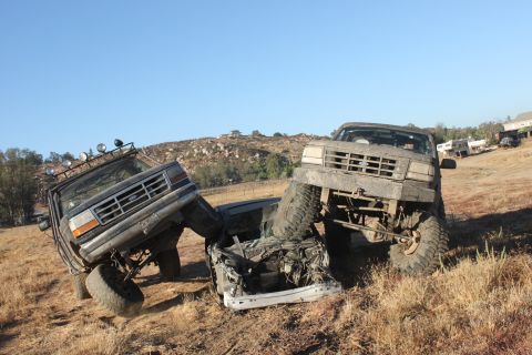 Two mud-covered 1992 Ford Broncos are crashed together on a dry, grassy hill under a clear blue sky
