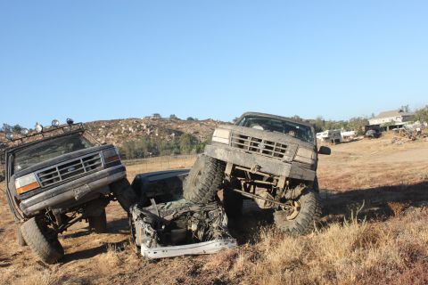 Two lifted trucks are on top of a crushed car in a dry, grassy field. One truck is dark, the other is lighter