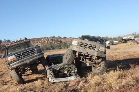 Two 1992 Ford Broncos are off-roading, one is on top of a crushed car